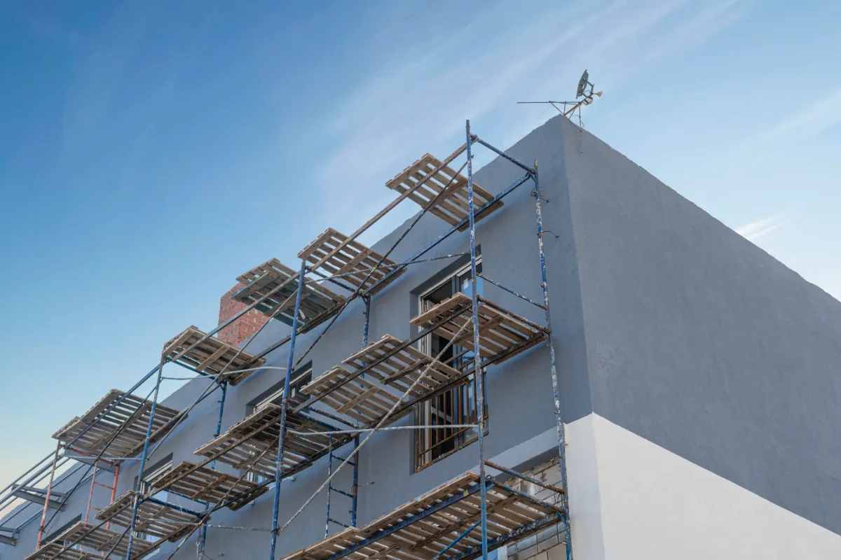 Scaffolding surrounds a blue-gray building under construction, with wooden planks forming platforms against a clear sky.