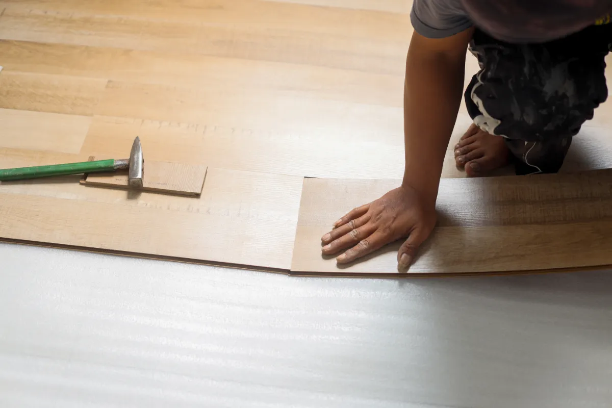 Worker lays wooden floorboards, pressing a plank into place as a hammer rests nearby on the boards