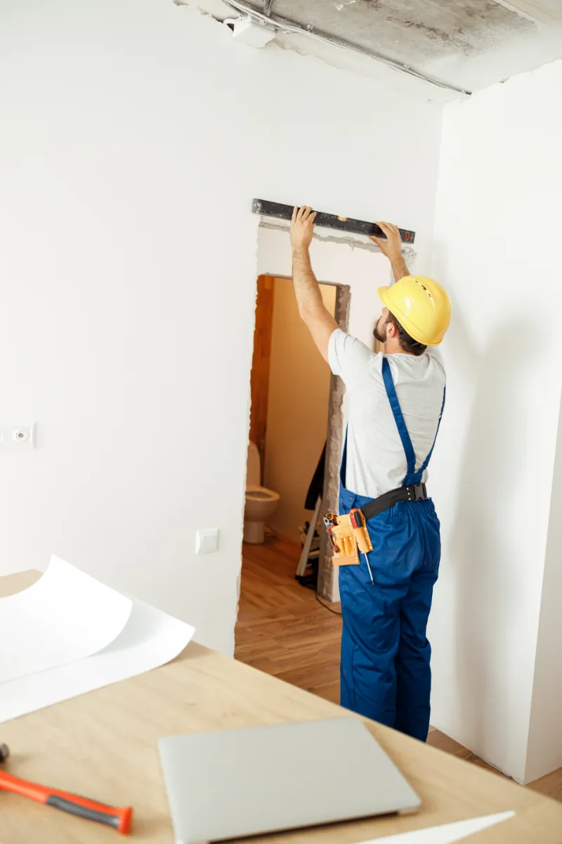 Construction worker in a yellow hard hat aligns a door opening with a level during renovation work on a white wall.