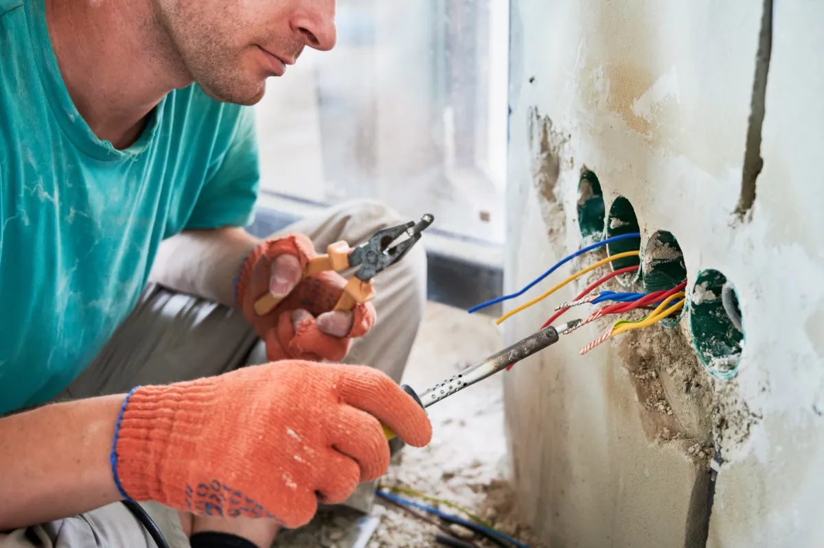 Electrician wearing orange gloves connects colorful wires into a wall electrical box during installation.