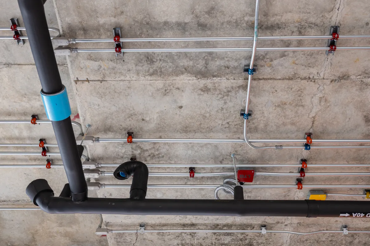 Industrial ceiling with silver metal pipes, blue clamps, and orange/red fittings mounted on concrete wall/ceiling. A large black insulated pipe runs across the foreground.