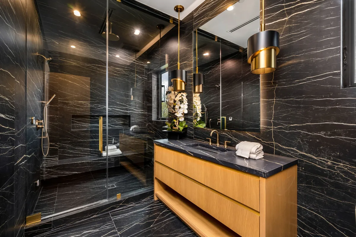 Luxurious black marble bathroom with gold fixtures, glass shower, and a wooden vanity with a black countertop connotes a high-end, spa-like space.