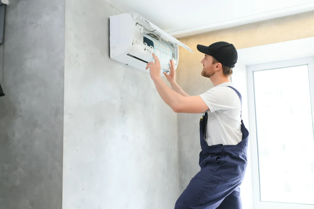 HVAC technician in a white shirt and blue overalls services a wall-mounted air conditioner on a concrete wall.