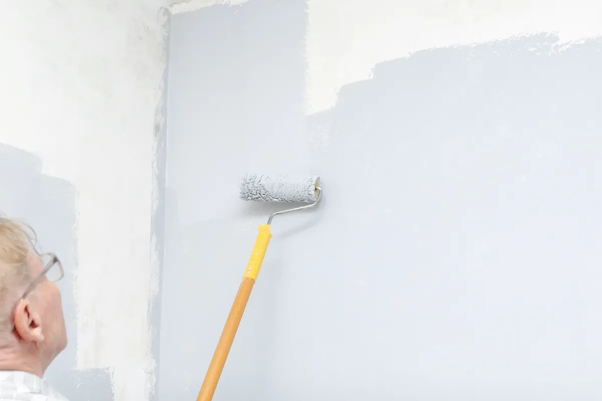 Person wearing glasses rolls light-gray paint onto a wall with a paint roller attached to a wooden handle nearby edge of room visible on left.