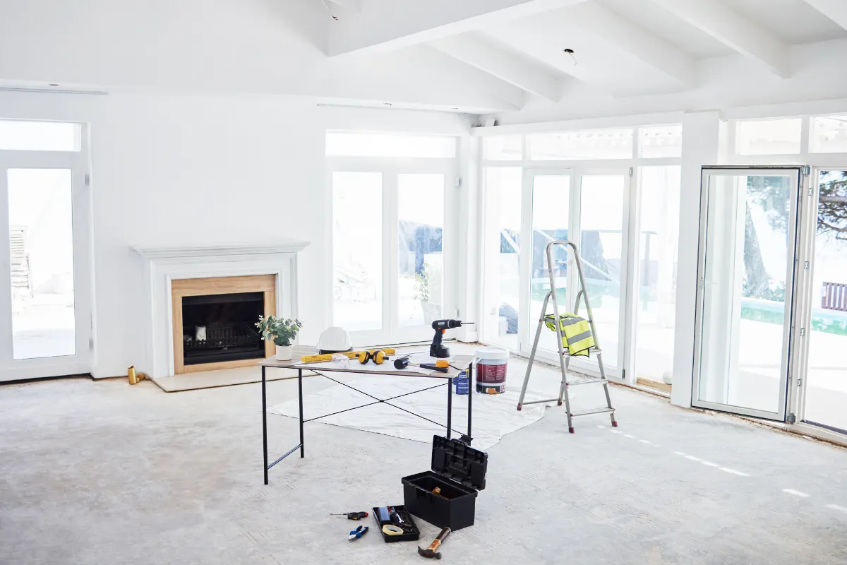 A bright living room under renovation with a fireplace, large windows, and a ladder; tools and paint supplies spread on a black table and floor.