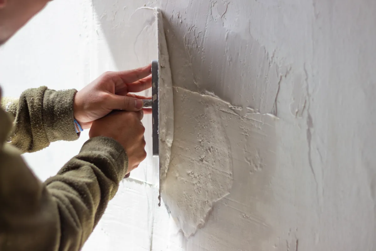 Person smoothing plaster onto a wall using a metal trowel, close up of hands and wall surface.