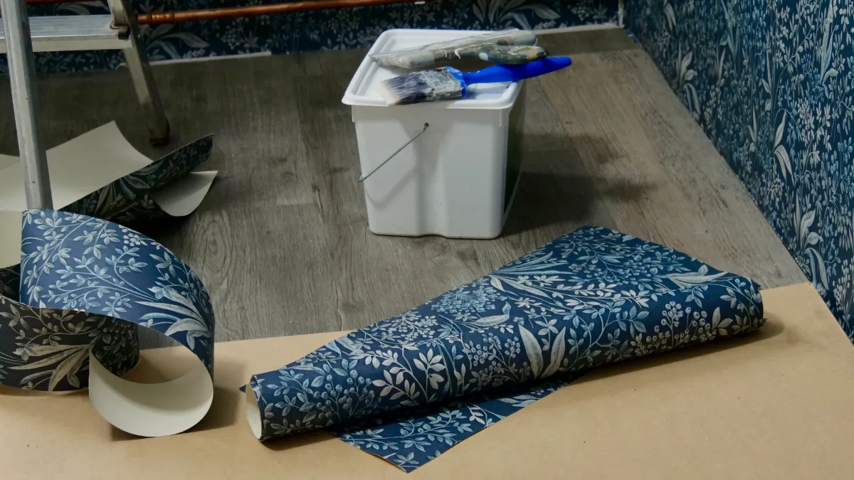 Rolls of blue floral wallpaper spread on the floor next to a white bucket filled with brushes, in a room with matching wallpapered walls and scraps nearby.