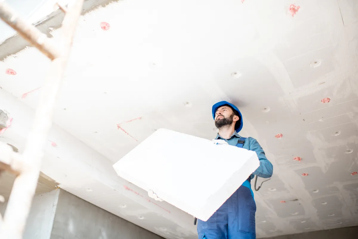 Construction worker wearing a blue helmet and overalls lifts a large white foam block in a building under construction