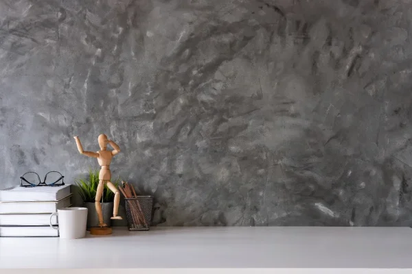 Wooden artist mannequin posing with one arm raised on a white desk beside a mug, books, and a pencil cup against a gray textured wall.