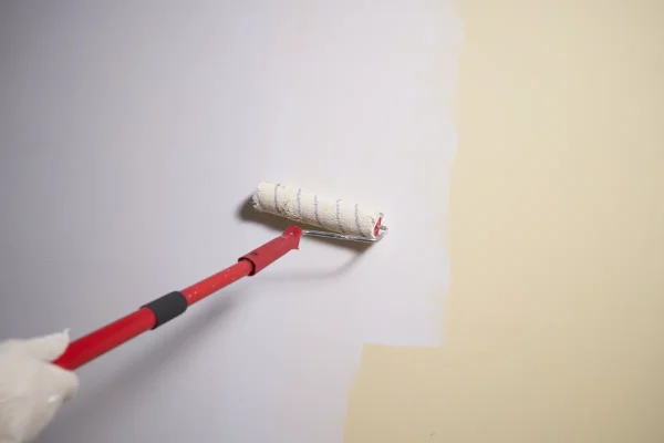 Gloved hand operates a paint roller applying white paint to a beige wall far background shows edge of color.